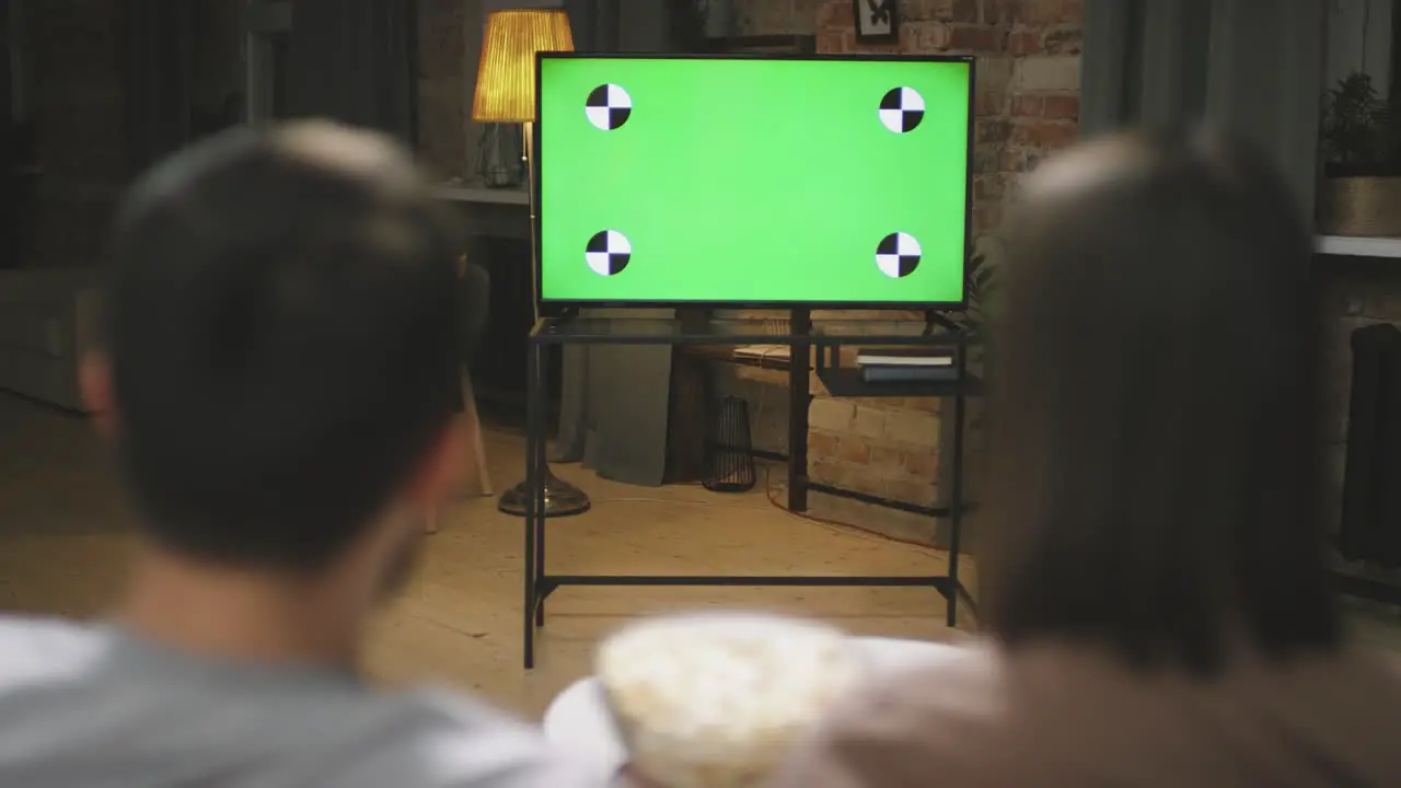 Rear View Of A Couple Sitting On The Couch Looking At A Screen While Eating Popcorn