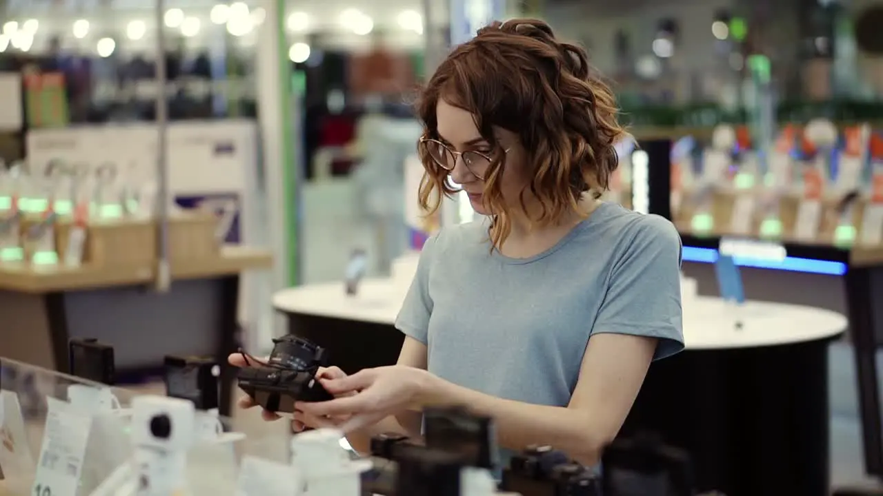 Young Curly Woman In Glasses Shopping For A New Photocamera In The Electronics Store