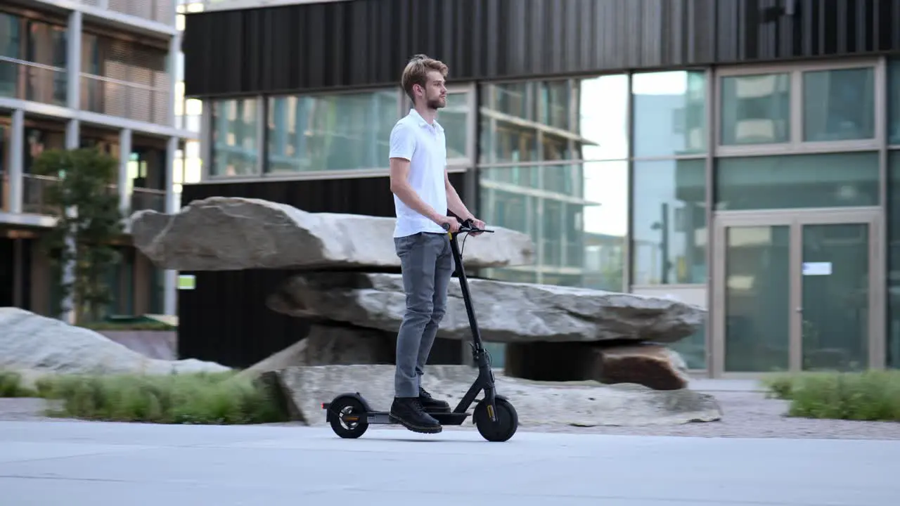 Businessman rides past modern building on his e-scooter wide shot