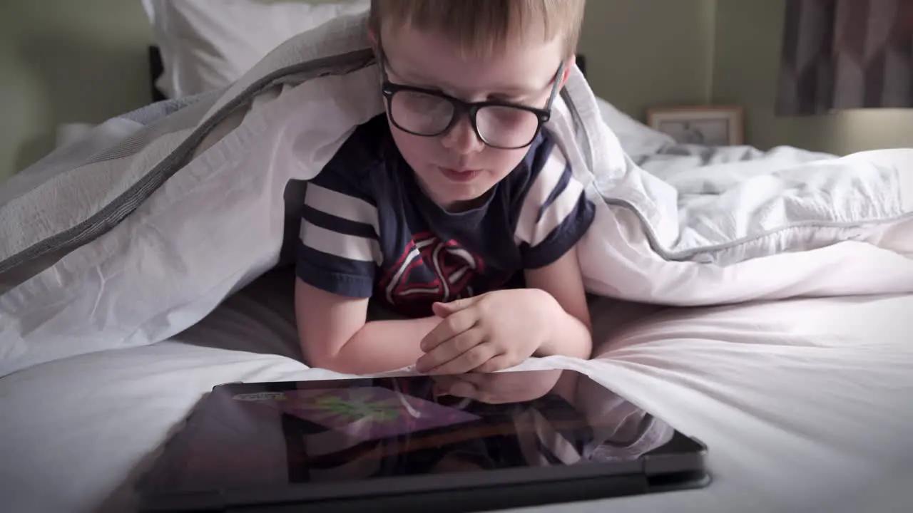 A six years old boy reading a computer tablet while lying in his bed