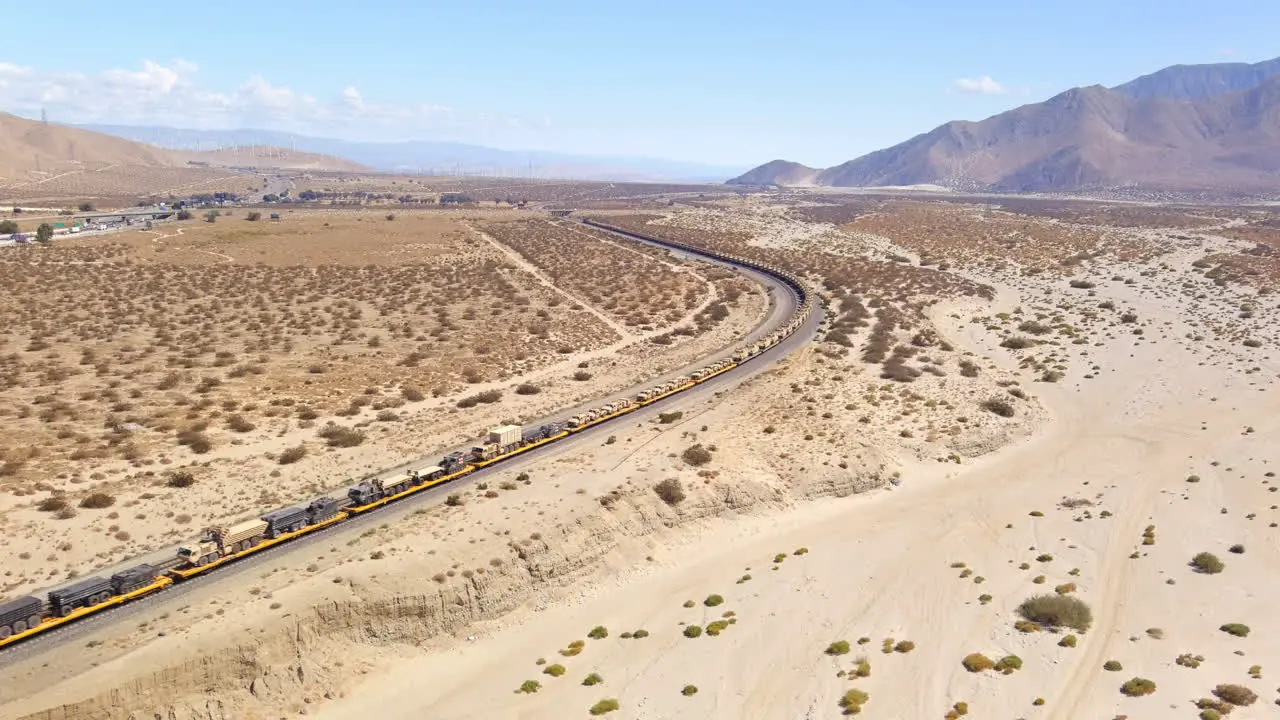 Rising wide view aerial of American Army train moving combat vehicles through the desert in California