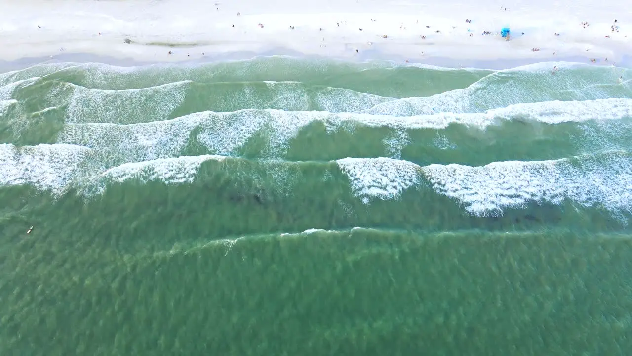 Birdseye drone clip of green water waves rolling onto white sand beach with people sunbathing below