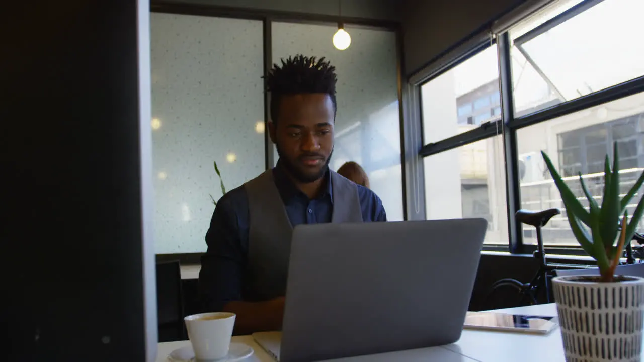 Front view of young black businessman working on laptop with a cup of coffee sitting at desk 4k