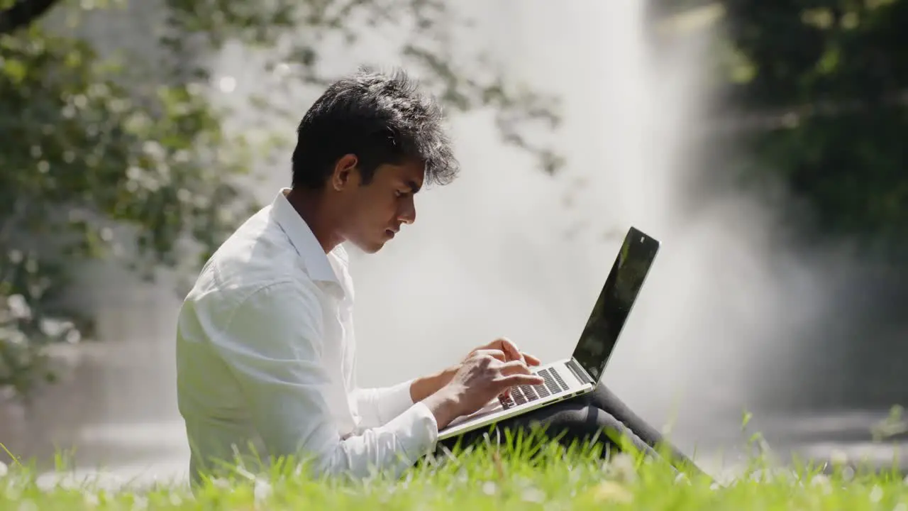 Young professional with laptop working remotely from public park Water sprays from fountain in Background