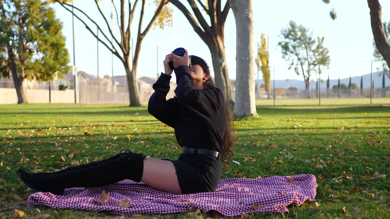 A beautiful millennial girl photographer taking pictures with a digital camera on a blanket in a park surrounded by autumn leaves