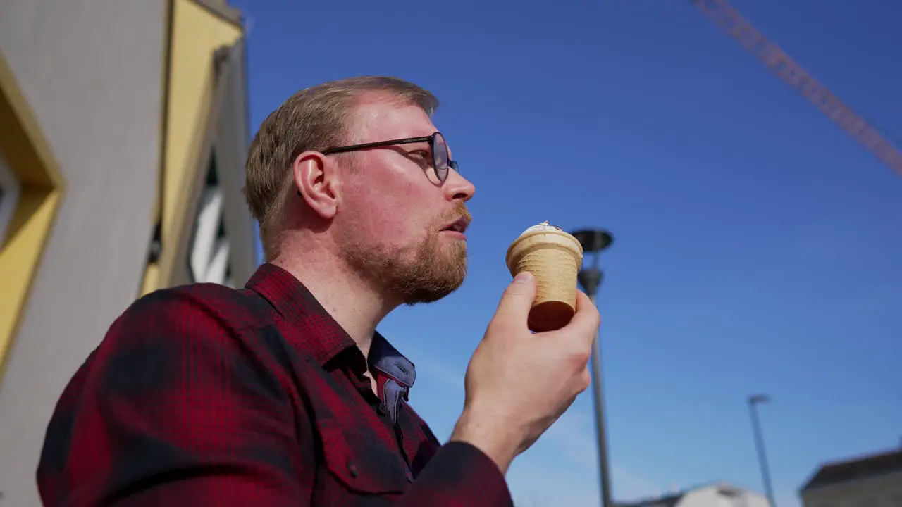 Young Man with Beard and Glasses Eats Ice Cream Outside on Sunny Day Low Angle