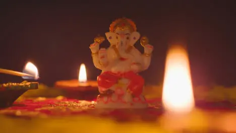 Hand Lighting Diya Lamps Around Statue Of Ganesh On Table Decorated For Celebrating Festival Of Diwali