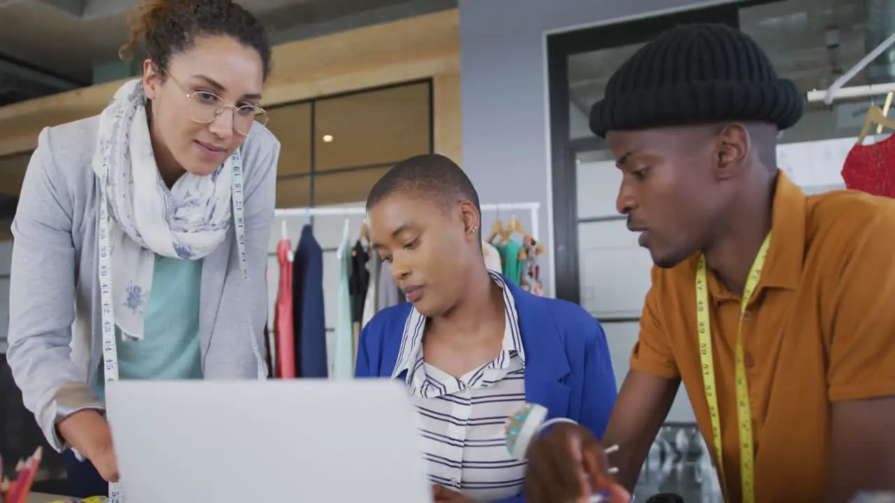 Diverse female and male designers standing in discussion using laptop in fashion studio