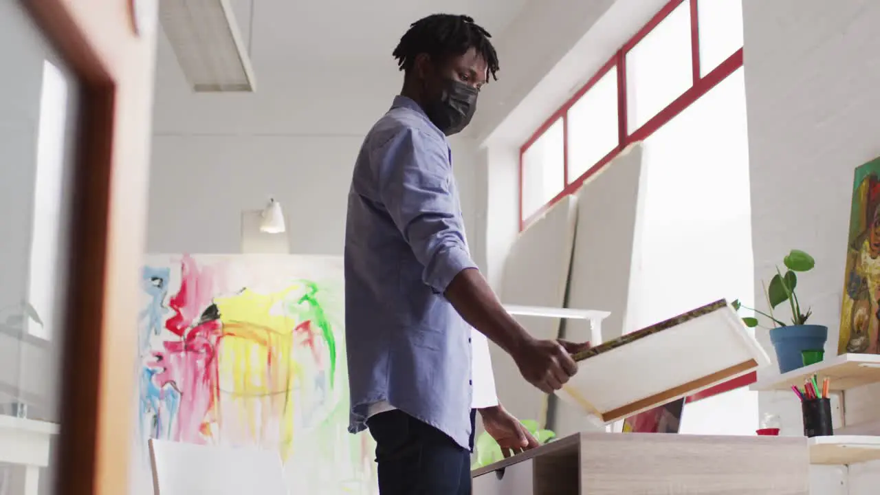 African american male artist wearing face mask holding a painting and using laptop at art studio