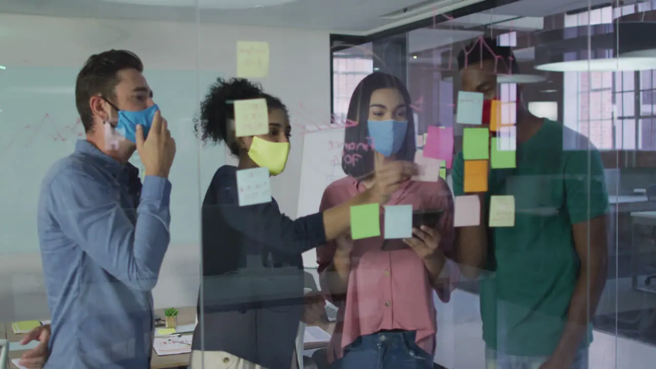 Diverse colleagues wearing face masks using memo notes on glass wall having a discussion