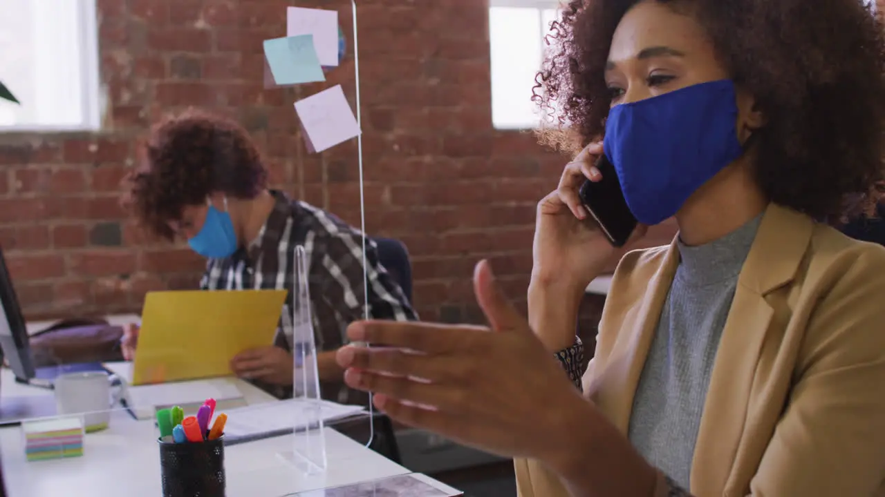 Mixed race businesswoman talking on phone sitting in front of computer wearing face mask