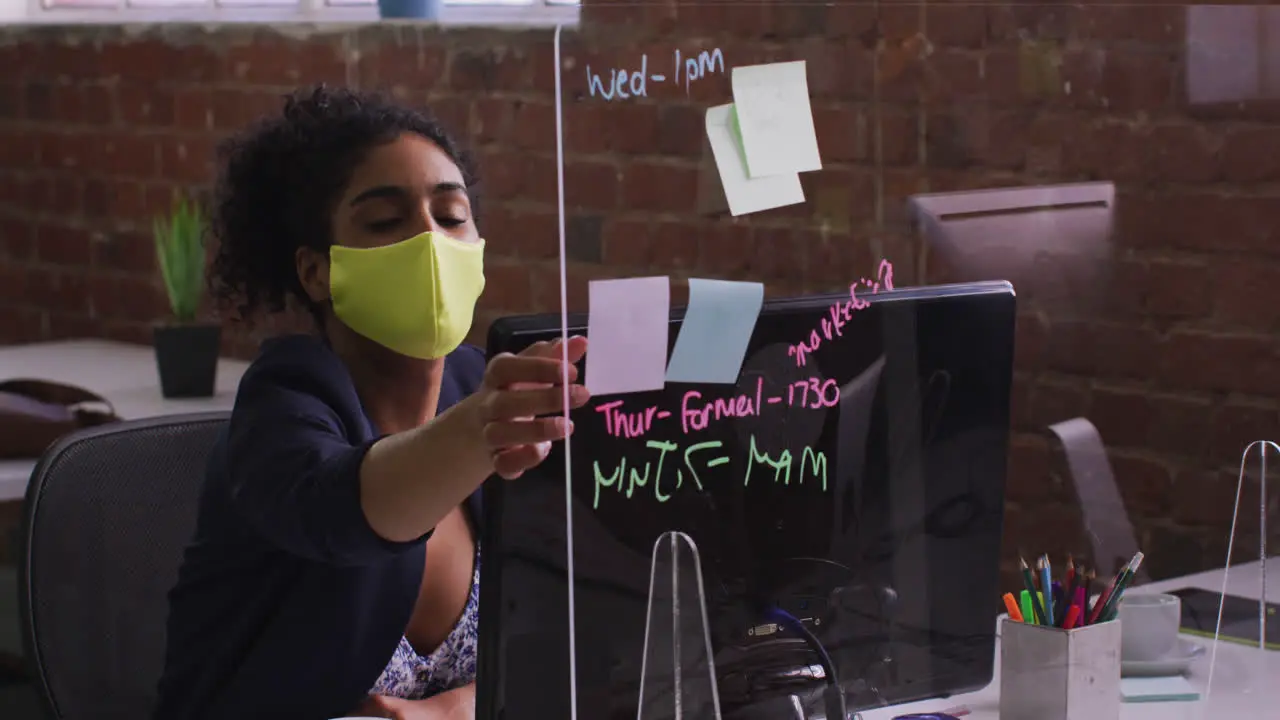 Mixed race businesswoman using memo notes sitting in front of computer wearing face mask