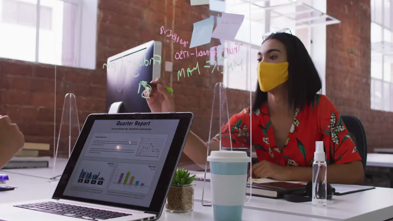 Mixed race businesswoman writing on sneez screen sitting in front of computer wearing face mask