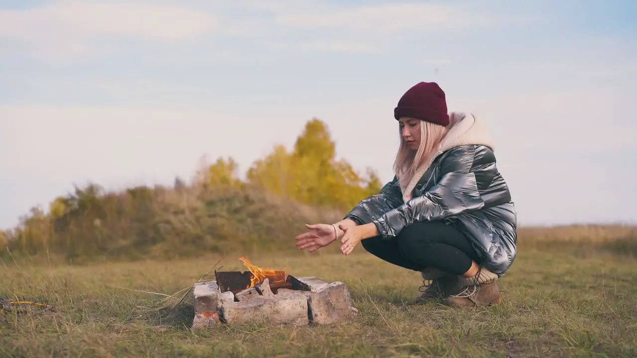 woman hiker heats hands at bonfire in camp in autumn