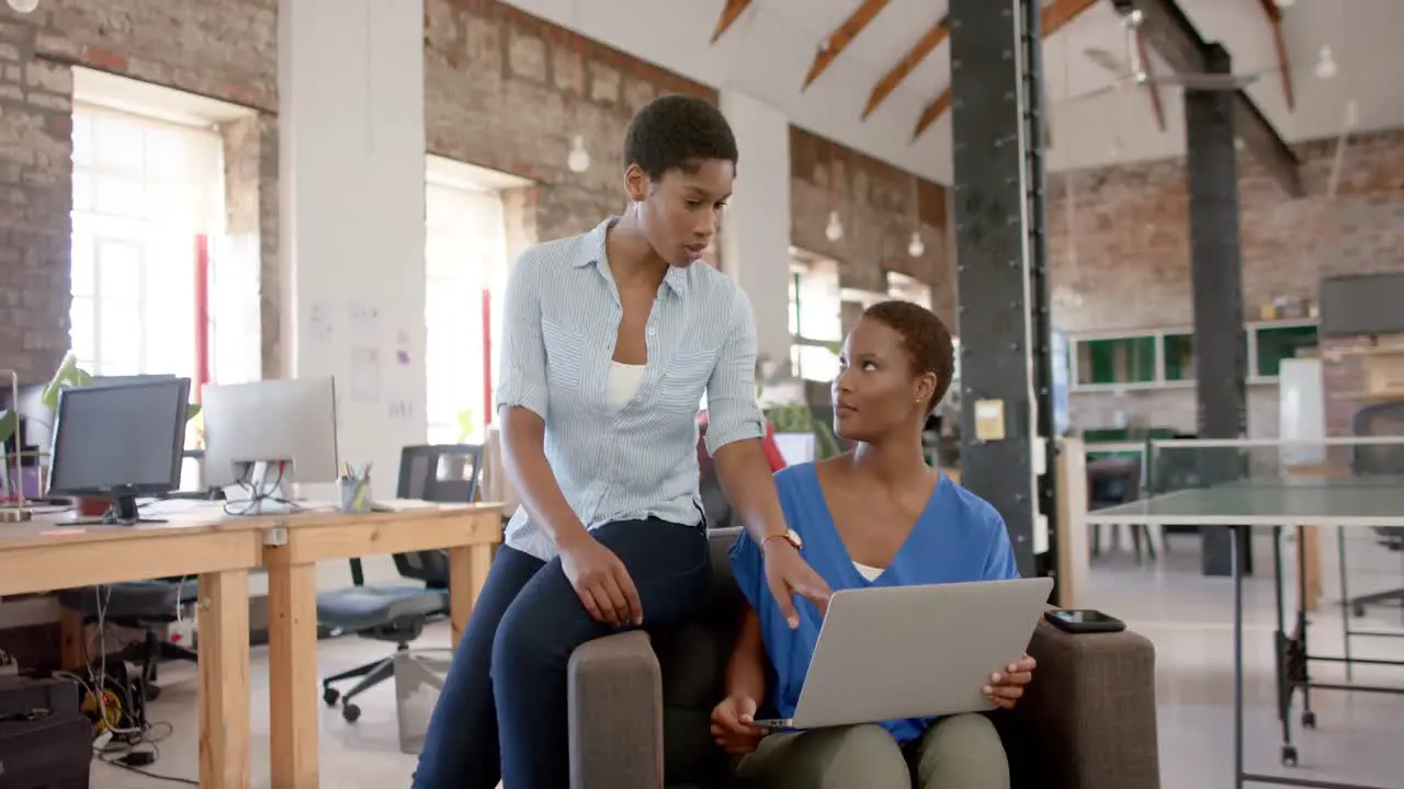 African american female colleagues using laptop and talking in creative office slow motion