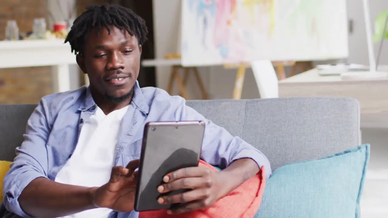 African american male artist smiling while using digital tablet sitting on the couch at art studio