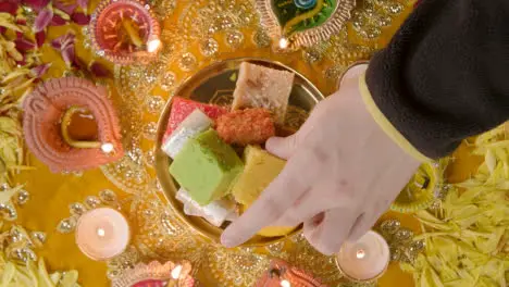 Overhead Shot Of Hand Picking Up Indian Sweets In Bowl On Table Decorated To Celebrate Festival Of Diwali