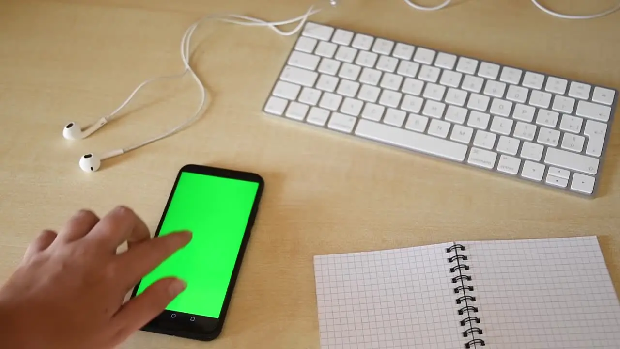 beautiful female hand scrolling and tapping on phone with green screen on a minimal workplace with keyboard earphones and notebook