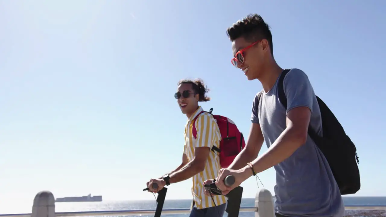 Happy diverse gay male couple using scooters at promenade by the sea slow motion