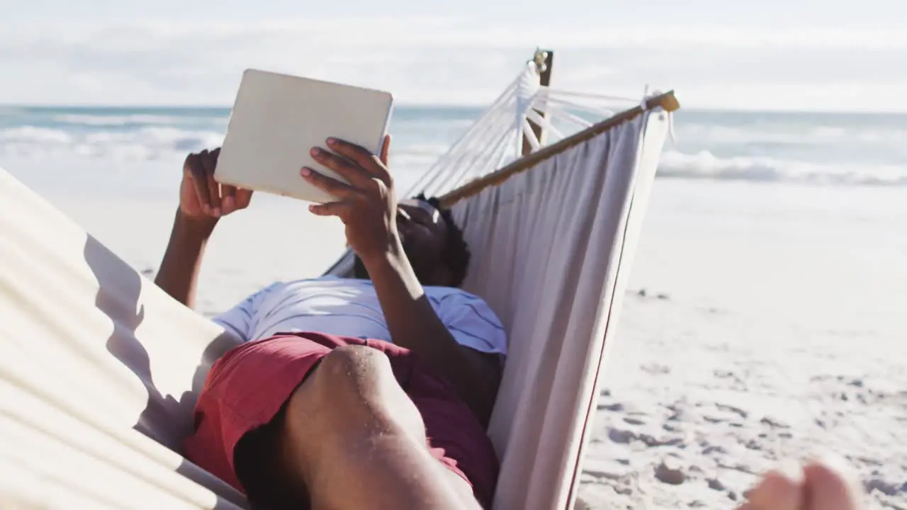 African american man using digital tablet while lying on a hammock at the beach