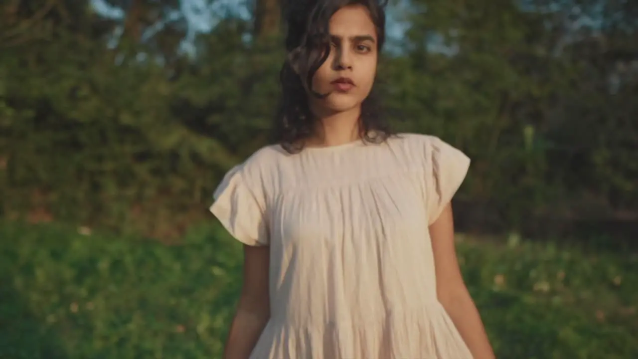 Slow motion backward dolly shot of a young charming indian woman in a dress looking at the camera with a serious look while she is in nature