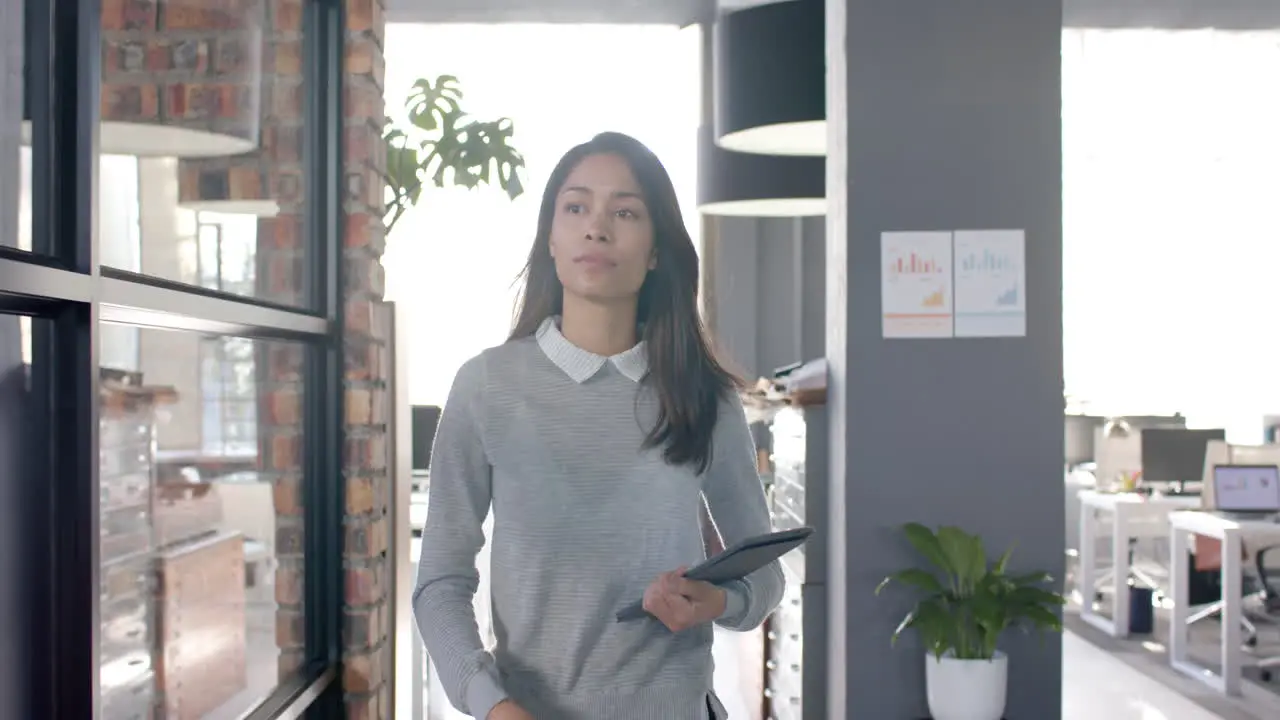 Biracial casual businesswoman walking through office with tablet slow motion