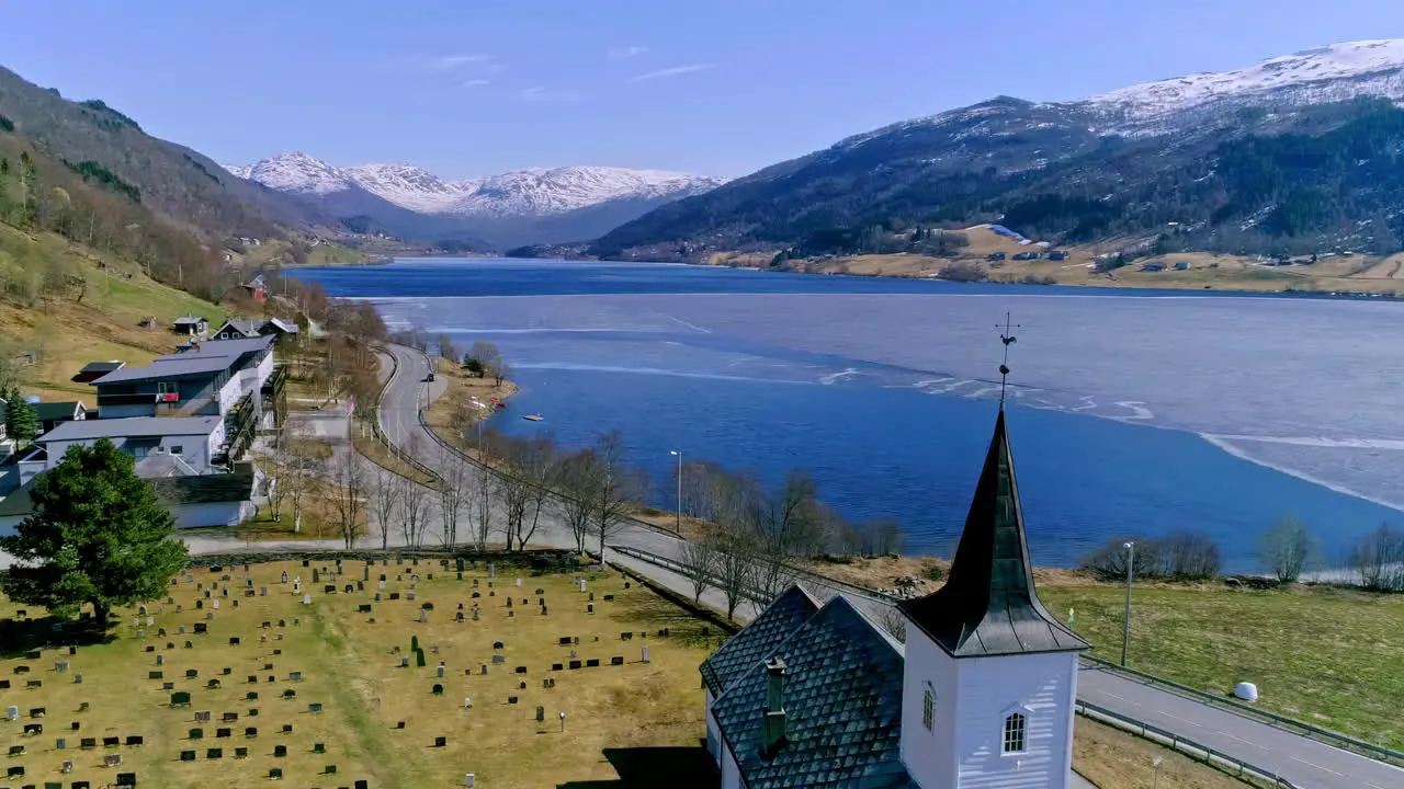 Aerial shot of a snowy lake next to a green valley with a church with a weather vane and a cemetery