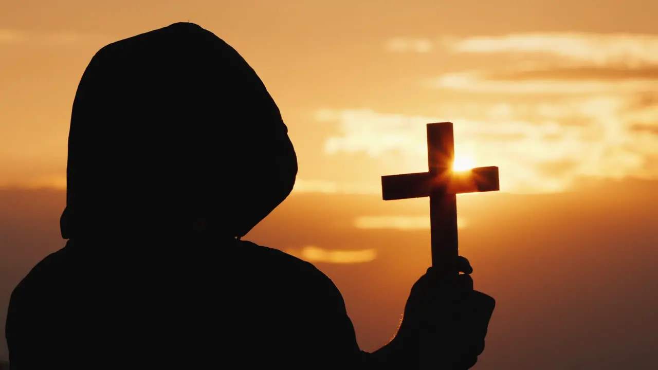 A Monk In A Hood With A Crucifix In His Hands Stands Against The Backdrop Of A Dramatic Sky At Sunse