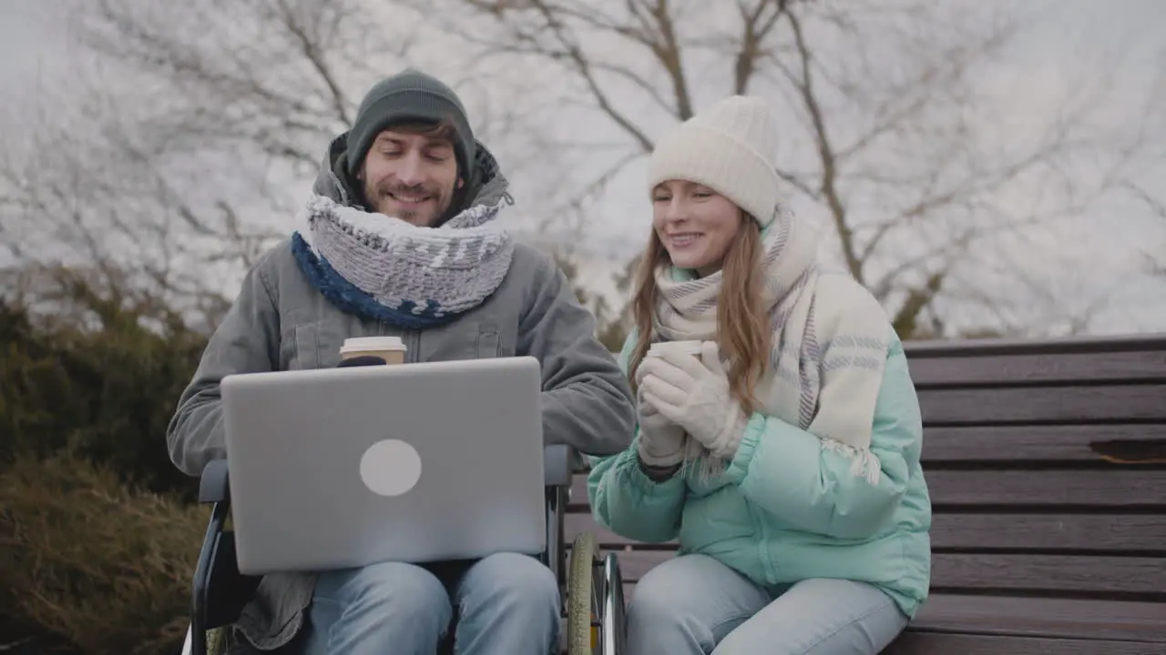 Disabled Man In Wheelchair And His Friend Watching Something Funny On Laptop Computer While Drinking Takeaway Coffee At Urban Park In Winter 3