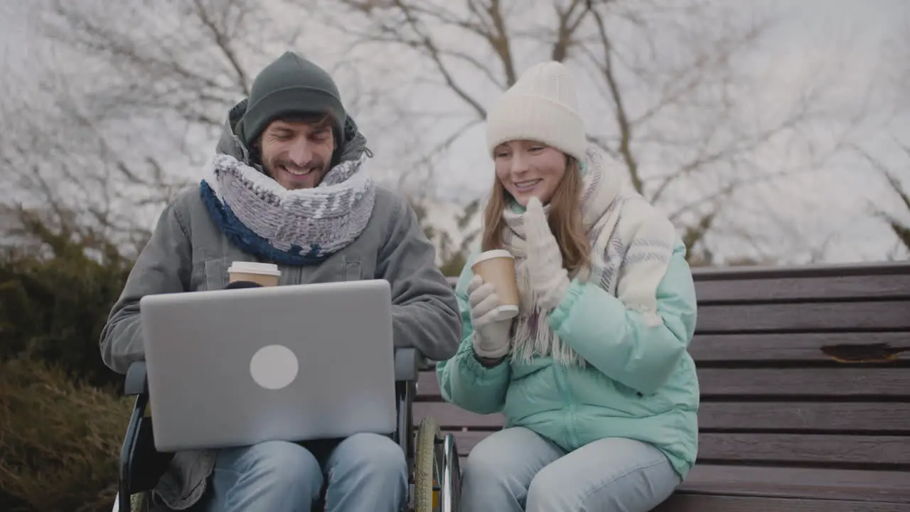 Disabled Man In Wheelchair And His Friend Watching Something Funny On Laptop Computer And Laughing Together At Urban Park In Winter