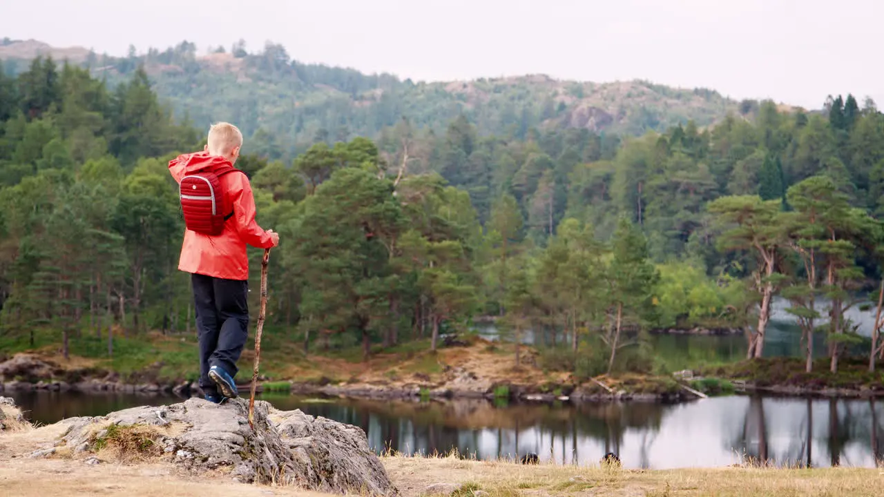 A boy balancing on a rock by a lake admiring the view on the left hand side of frame back view Lake District UK