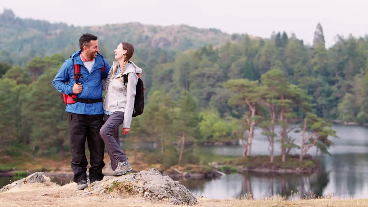 Young adult couple standing on the left of shot against a lake view laughing to camera Lake District UK