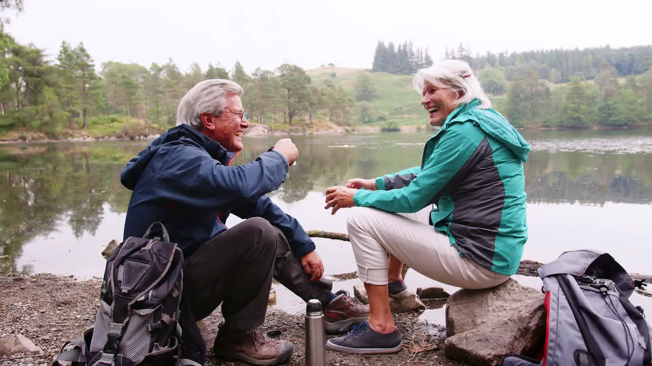 Senior couple sitting on rocks by a lake laughing and talking during a camping holiday Lake District UK