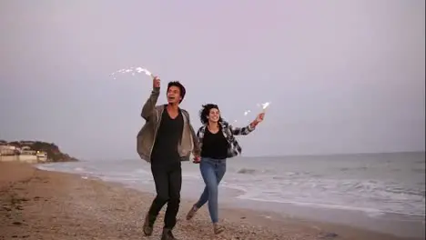 Young happy multi ethnic couple holding burning sparkling candles and running by the sea during sunset Slow Motion shot