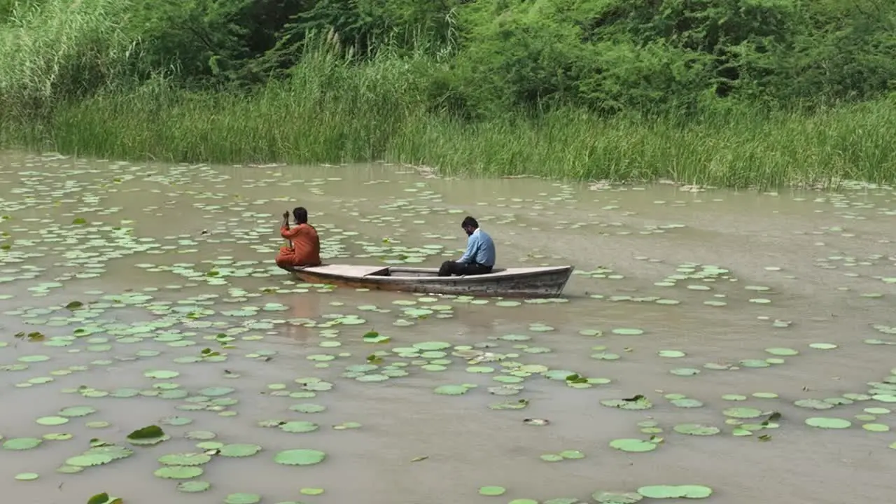 Aerial Orbit Motion Around Two Men On Small Wooden Boat On Botar Lake With Small Green Lily Pads Floating On Surface