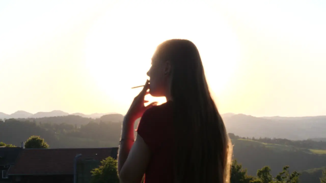 Young woman standing outside smoking cigarette and enjoying the view of landscape with sunset