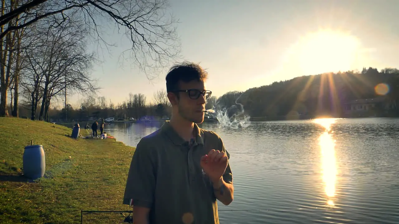 Man standing beside a lake smoking as the sun sets in the distance