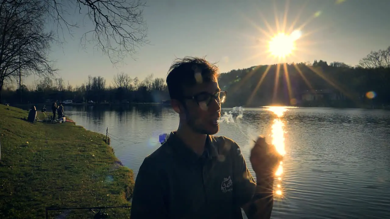 Man inhales deeply from a cigarette while standing beside a lake at sunset