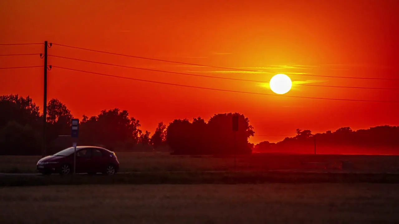 A car parked along a countryside road as a fiery sun descends behind a copes of trees time lapse