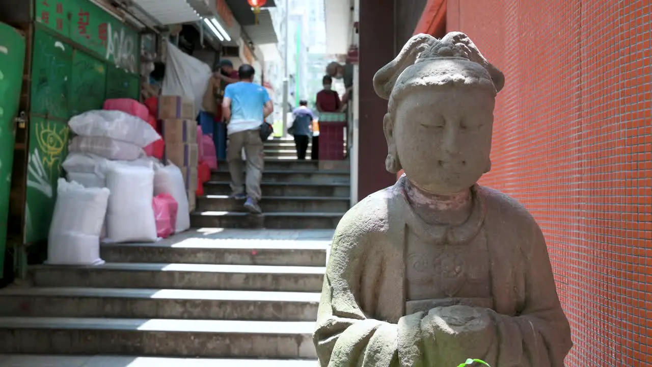 Pedestrians walk up the stairs on a narrow street as they pass a stone Buddha statue in a district known for its numerous antique shops