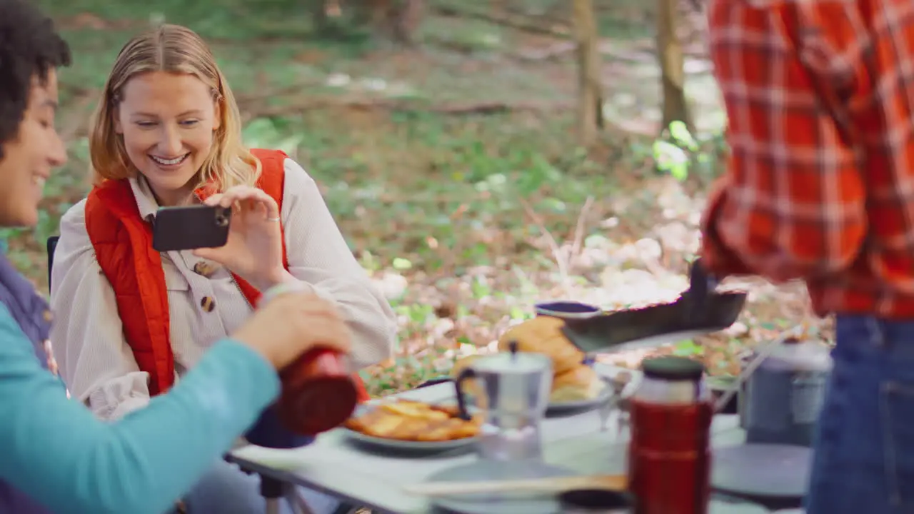 Woman Taking Photo Of Food On Mobile Phone As Female Friends On Camping Holiday Eat Meal By Tent