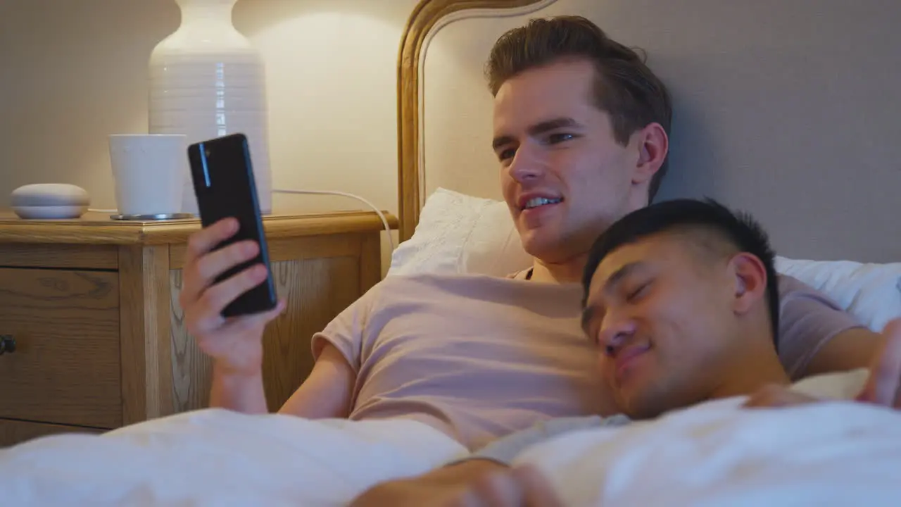 Loving Same Sex Male Couple Lying In Bed At Home Looking At Social Media On Mobile Phone