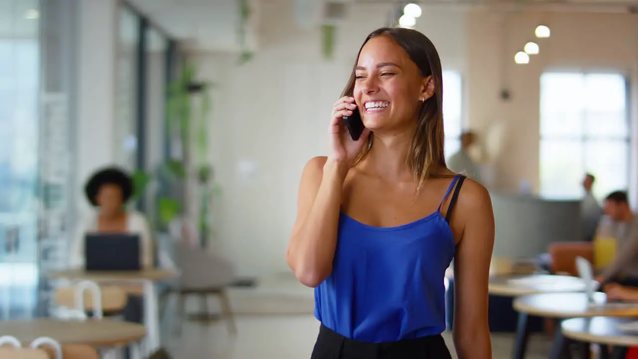 Young Smiling Businesswoman Standing In Busy Modern Open Plan Office Talking On Mobile Phone