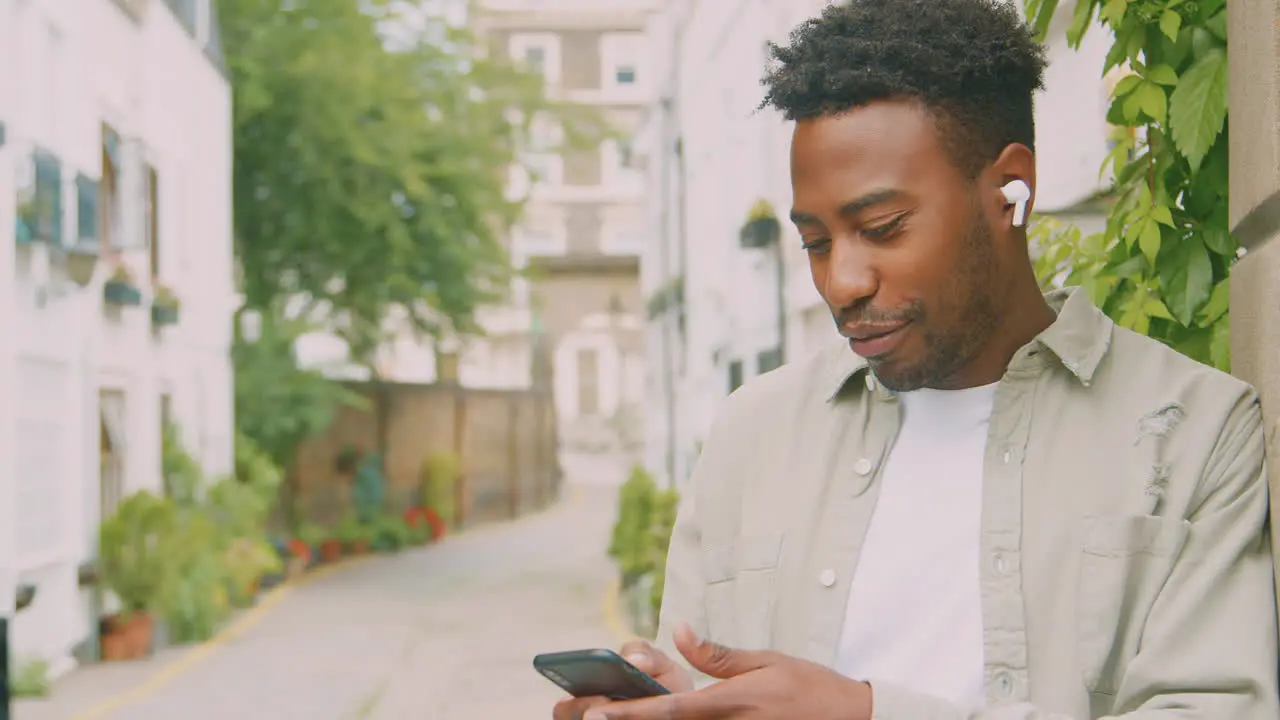 Young Man Travelling Through City Listening To Music On Wireless Earbuds On Mobile Phone