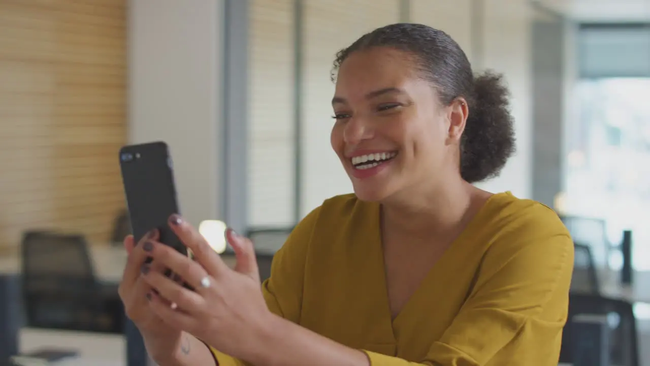 Young Businesswoman Having Video Chat On Mobile Phone Call Standing In Modern Open Plan Office