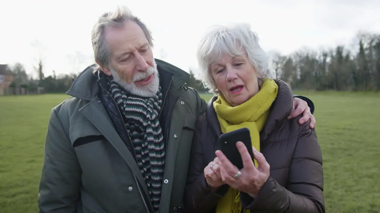 Senior Couple Looking At Mobile Phone On Autumn Or Winter Walk Through Park Together