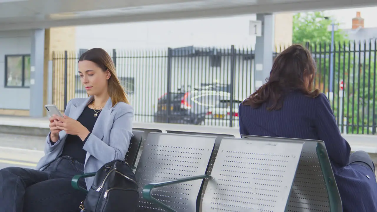 Two Businesswomen Commuting To Work Waiting For Train On Station Platform Looking At Mobile Phones