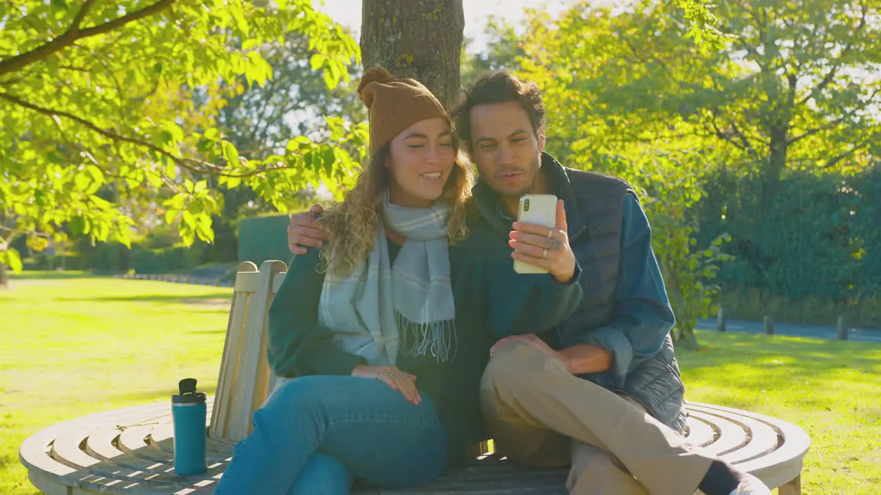 Couple Sitting On Bench In Autumn Park Making Video Call On Mobile Phone