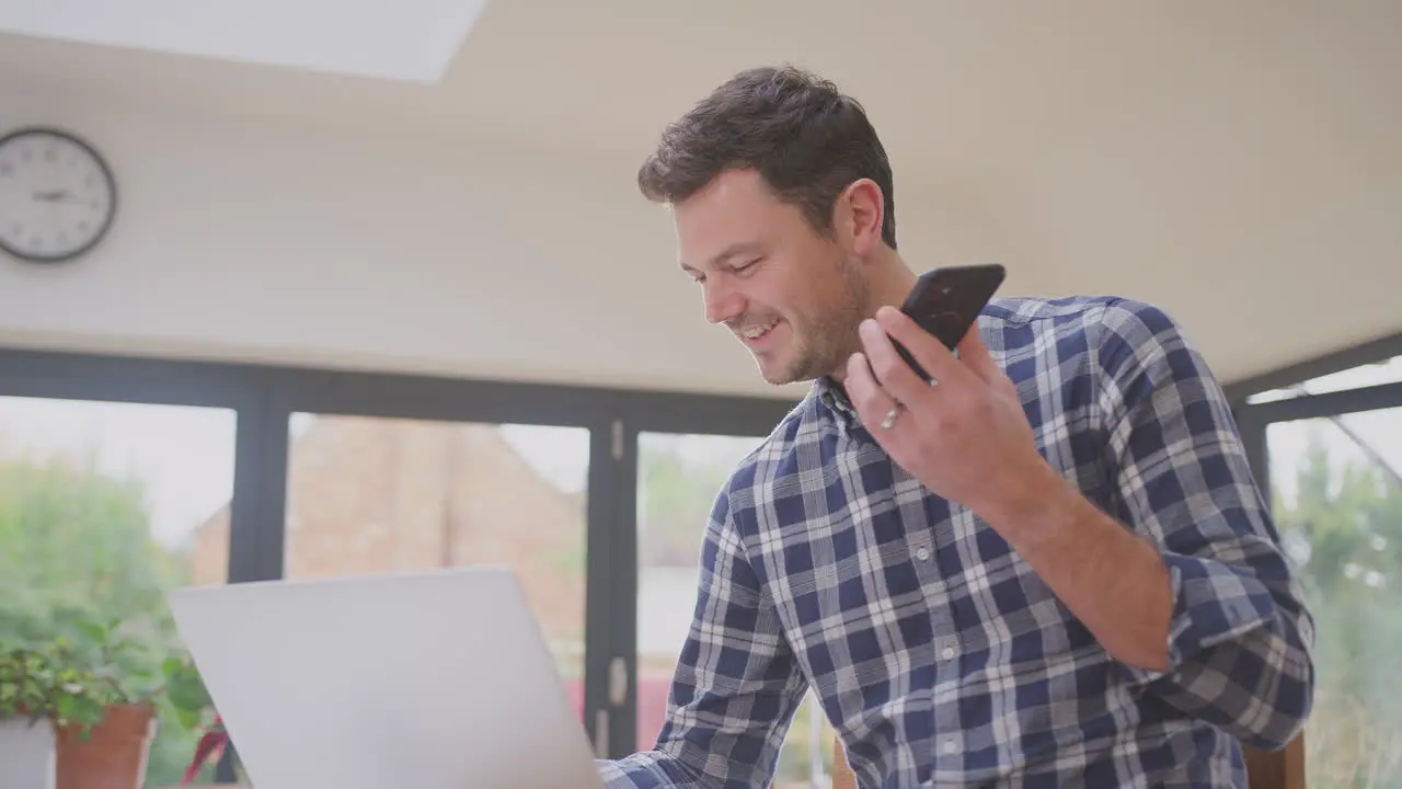 Man working from home using laptop on kitchen counter talking into microphone of mobile phone- shot in slow motion