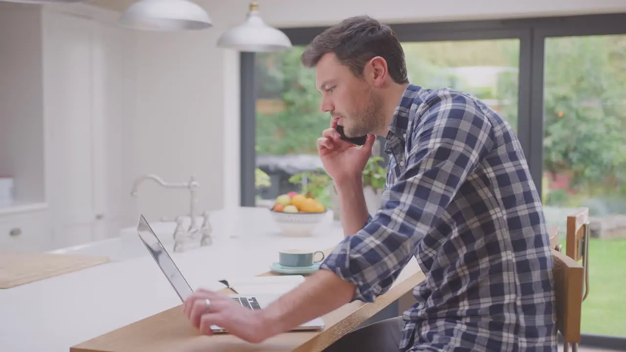 Man working from home using laptop on kitchen counter talking on mobile phone- shot in slow motion